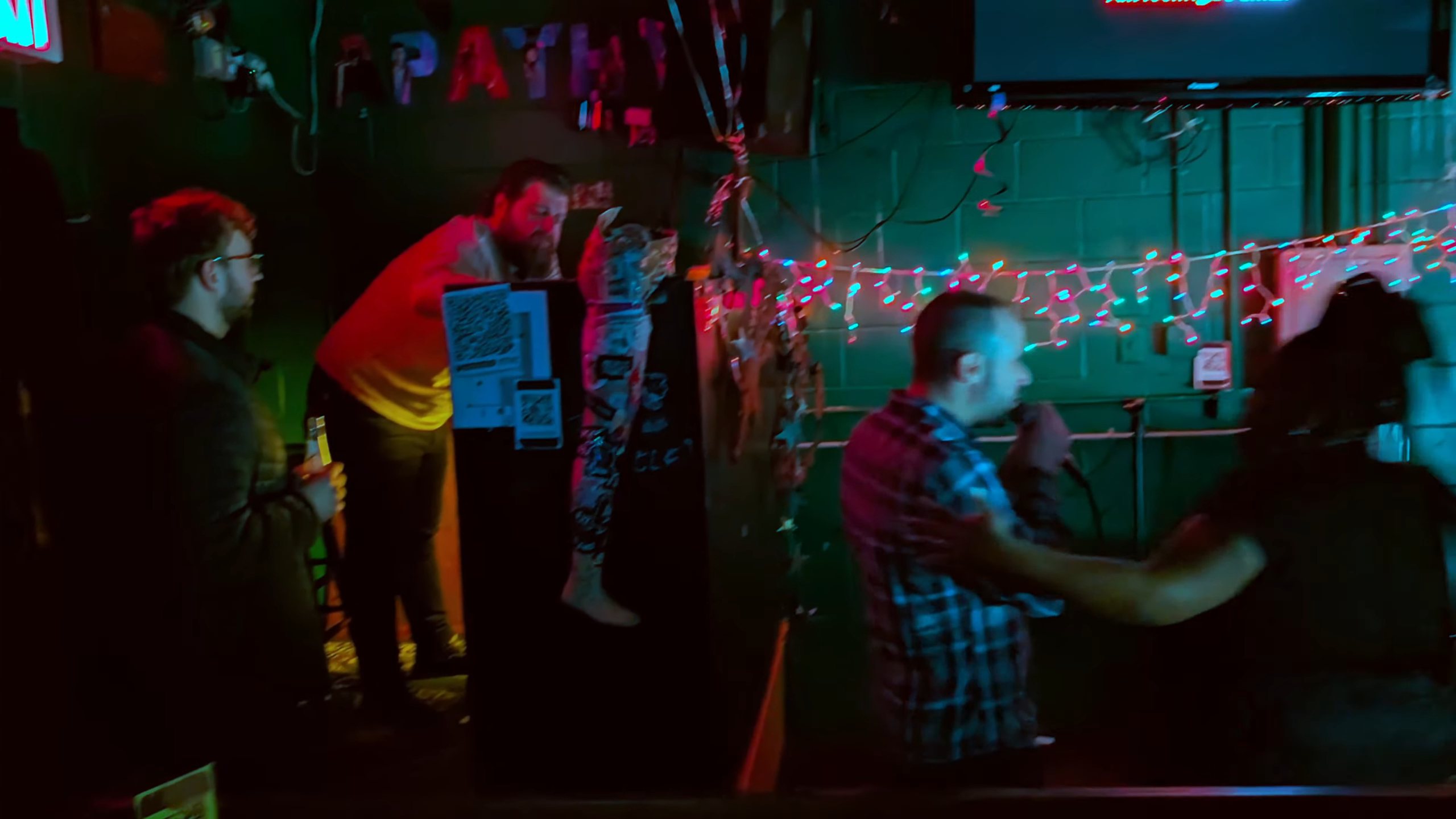 Dimly lit bar scene at Alligator Lounge with people enjoying karaoke, beer, and eclectic decor including string lights and stickers