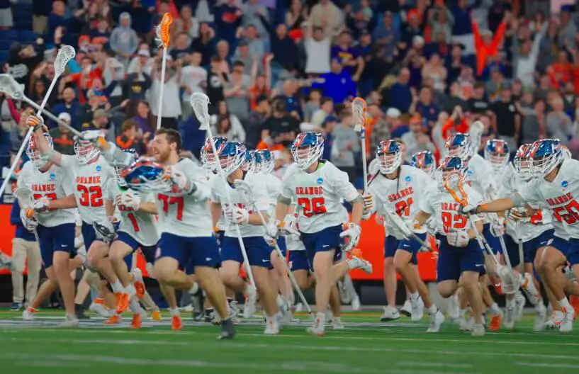 Orange team celebrates a victory, running onto the field with their lacrosse sticks raised high