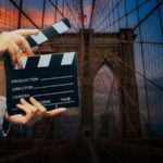 A person holds a film clapperboard in front of the Brooklyn Bridge at dusk, highlighting classic movies set in Brooklyn