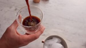 A person pours freshly brewed espresso into a clear glass cup next to a container of ice on a marble counter