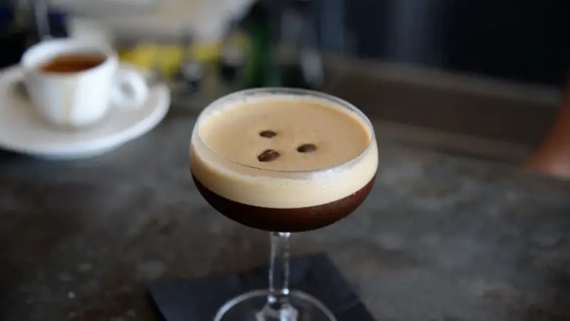 A freshly made espresso martini in a coupe glass topped with three coffee beans sits on a dark napkin on a bar counter