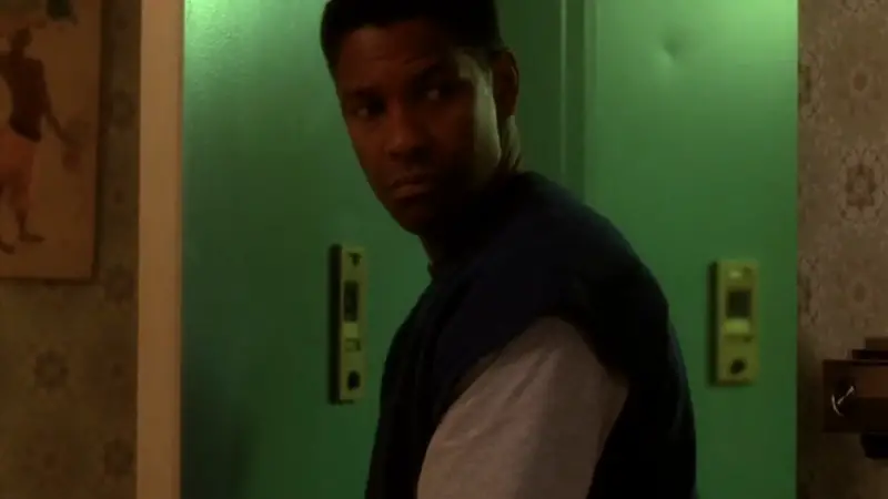 A man stands by a green door, looking back with a serious expression in a dimly lit Brooklyn apartment