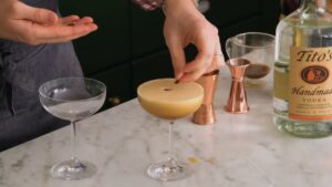 A person places coffee beans on top of a freshly poured espresso martini in a coupe glass next to an empty glass and cocktail tools