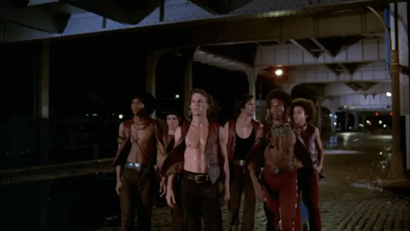 A gang of young men walk together under an overpass at night on wet Brooklyn streets