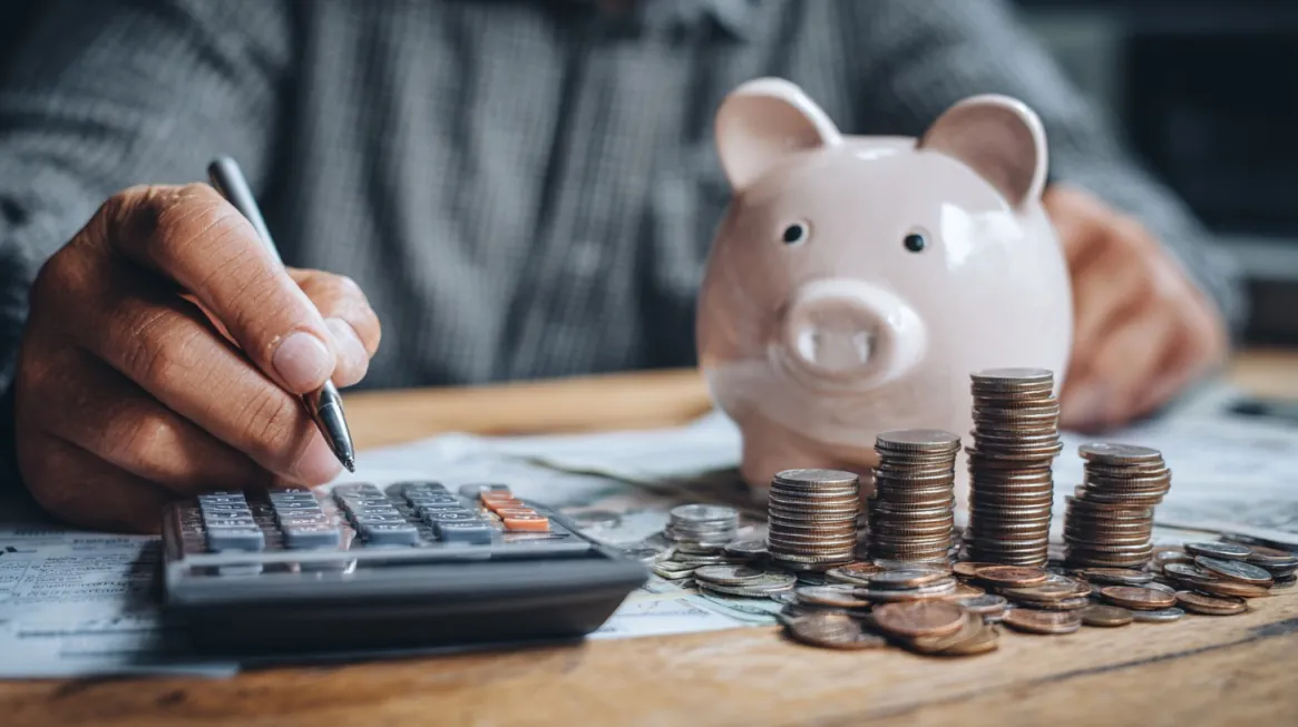 Close-up of a person using a calculator with a piggy bank and stacks of coins on the table