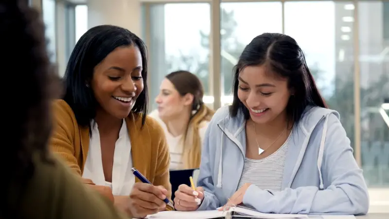 Two students study together at a table, smiling as they work on notes