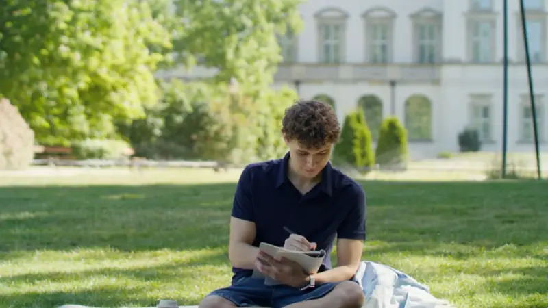 A student sits on the grass outside a university building, writing notes in a notebook