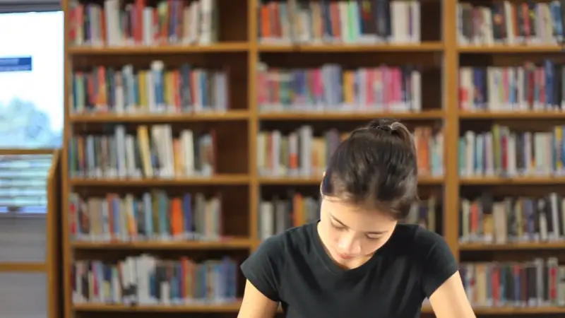 A student studies at a desk in a library surrounded by shelves of books