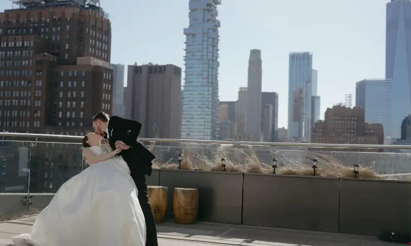A bride and groom share a kiss on a hotel rooftop, with the New York City skyline in the background