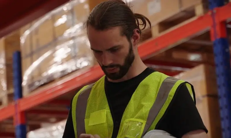 A man stands in a warehouse, holding a clipboard and reviewing inventory details