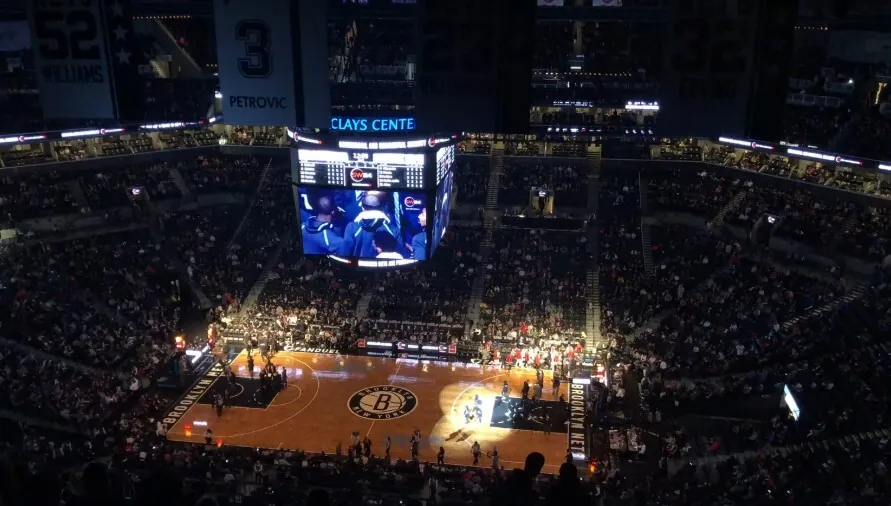 A packed basketball arena with illuminated court and large overhead scoreboard