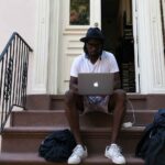 Person sitting on a brownstone stoop working on a laptop with bags and a charger nearby