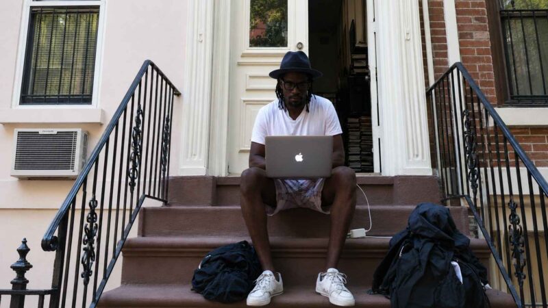 Person sitting on a brownstone stoop working on a laptop with bags and a charger nearby