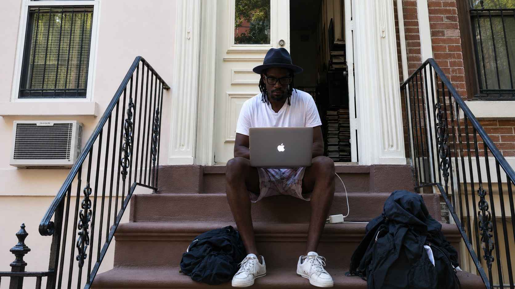 Person sitting on a brownstone stoop working on a laptop with bags and a charger nearby