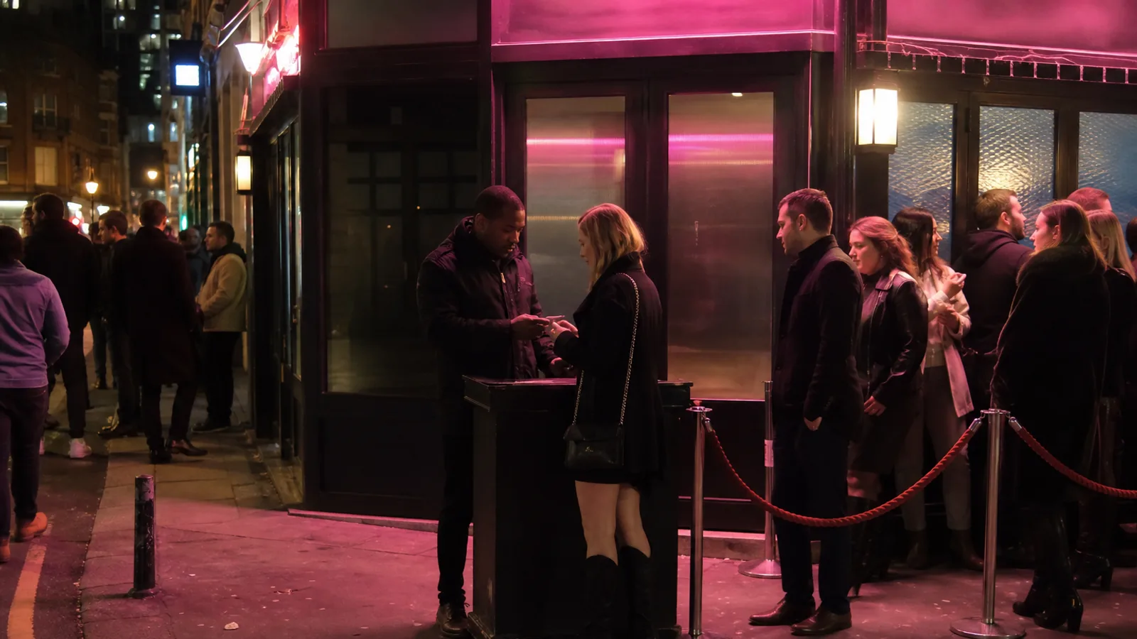A London venue door with a steward checking ID as a small queue waits under pink neon